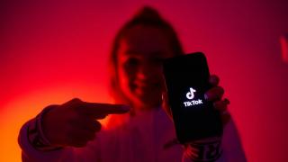 Ein Mädchen vor dunkelrotem Hintergrund mit einem Smartphone in der Hand, worauf "TikTok" zu lesen ist.