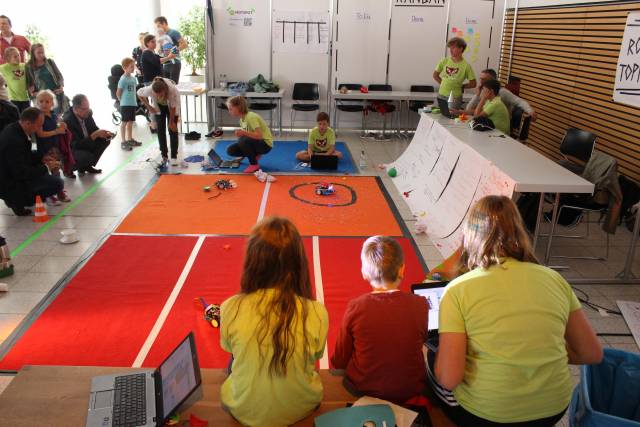Um einen roten Teppich sitzen Kinder und Erwachsene und schauen Robotern zu.
