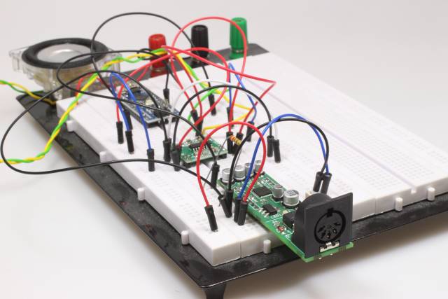 Ein großes weißes Steckbrett mit drei kleinen aufgesteckten Boards (Arduino nano, Verstärker und NanoSynth) die mit vielen Kabeln verbunden sind. Am Rand ist ein Lautsprecher zu erkennen.
