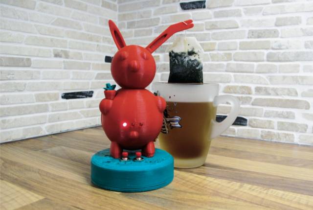 Ein roter Hase aus dem 3D-Drucker, dessen Ohr einen Teebeutel in einen Becher tunkt.