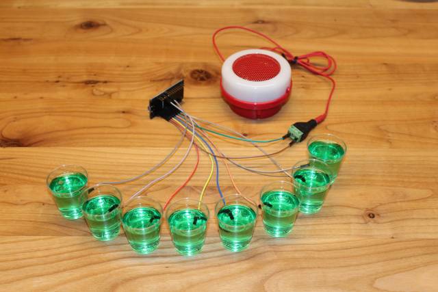 Acht Becher grüner Wackelpudding stehen auf einem Tisch. Sie sind über Kabel mit einem Board und dieses mit einem Lautsprecher verbunden.