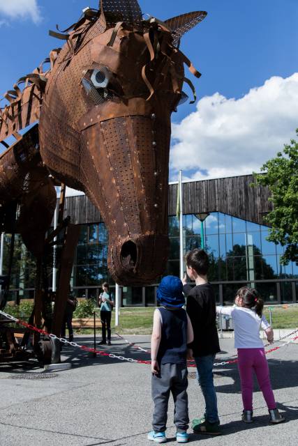 Drei Kinder stehen vor einem großen, dunklen Pferdekopf aus Metall.