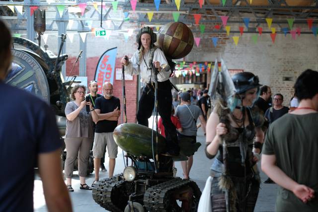Menschen in einer Halle, an der Decke hängen viele Fähnchen, in der Mitte eine Frau auf einem Gefährt mit kleinen Kettenrädern und hintem einem Ballon.