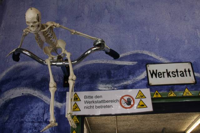 Neben einem Schild "Werkstatt" hängt an einer Wand ein Fahrradlenker, auf dem ein Skelett sitzt.