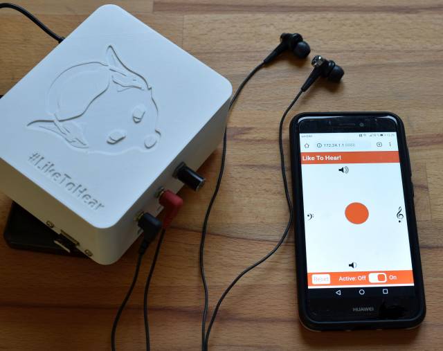 Eine weiß Box, daneben Kopfhörer und ein Handy mit offener liketohear-App.
