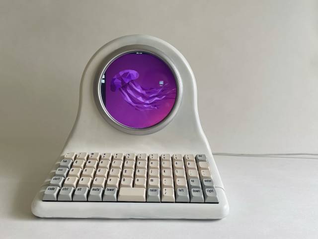 Weißer Computer mit rundem Bildschirm, auf dem eine lila Qualle zu sehen ist, und ortholinearer Tastatur.