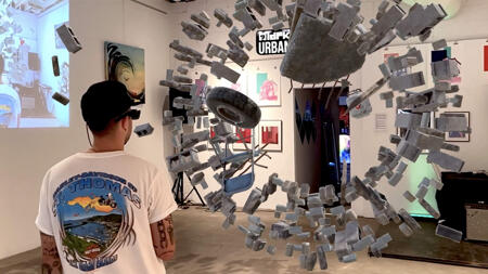 Ein Mann trägt ein Mixed-Reality-Headset und betrachtet in einer Galerie eine digitale 3D-Installation: Vor ihm schwebt ein holografischer Ring aus schwebenden Betonblöcken, einem Reifen und Metallteilen, die Teil einer Augmented-Reality-Kunstinstallation sind.