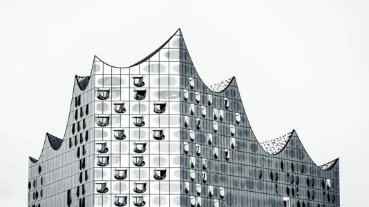 Modernes, gläsernes Gebäude mit wellenförmigem Dach und vielen geschwungenen Fenstern, aufgenommen vor hellem, neutralem Hintergrund.
