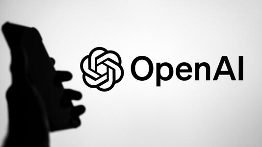OpenAI-Logo auf Bildschirm, davor Hand mit Smartphone in Silhouette