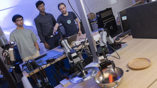 Einer von drei Forschern steuert mit Smartphone einen Roboterarm, der eine Karotte aus einer Metallschale greift. 