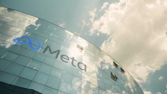 Meta-Logo auf der spiegelnden Glasfassade eines Bürogebäudes. Wolken und Sonnenlicht reflektieren sich in den Fenstern.