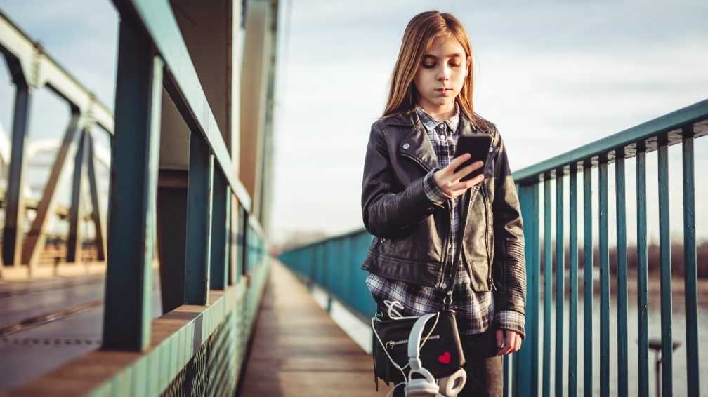 Ein Mädchen, das schwarze Lederjacke und Kopfhörer trägt, Smartphone verwendet und über eine Brücke geht