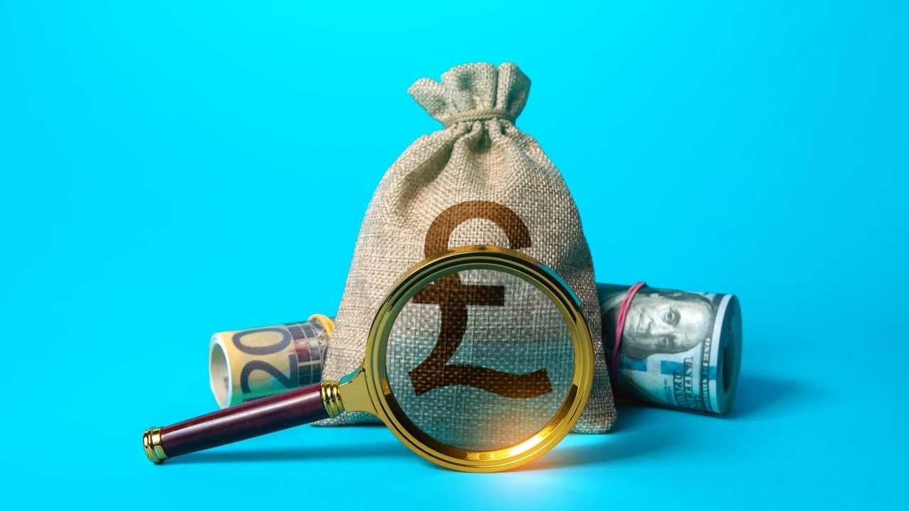 ein Geldsack voller britischer Pfund, daneben liegen eine Rolle US-Dollarscheine und Euro-Scheine. Vor diesem Arrangement ist eine Lupe.