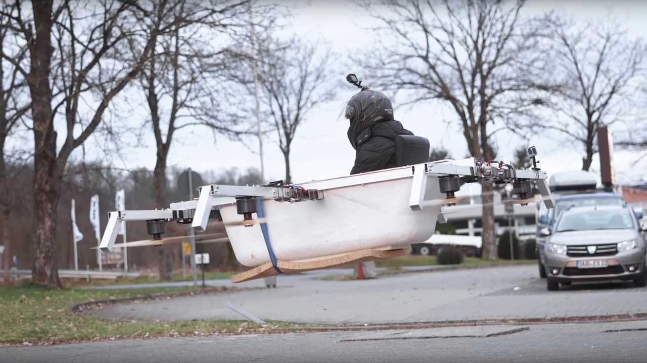 Eine weiße Badwanne schwebt über einem Parkplatz, an der Seite sind sechs Rotoren befestigt, in der Badewanne sitzt ein schwarz gekleideter Fahrer