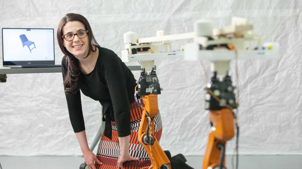 Eine junge Frau beugt sich neben einem Schreibtisch nach vorn und schaut zwei Roboterarme an, die ein Stück Holz halten
