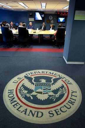 Vorderung: Siegel des Ministerium für Heimatsicherheit; Hintergrund: Leute sitzen an einem großen Konferenztisch