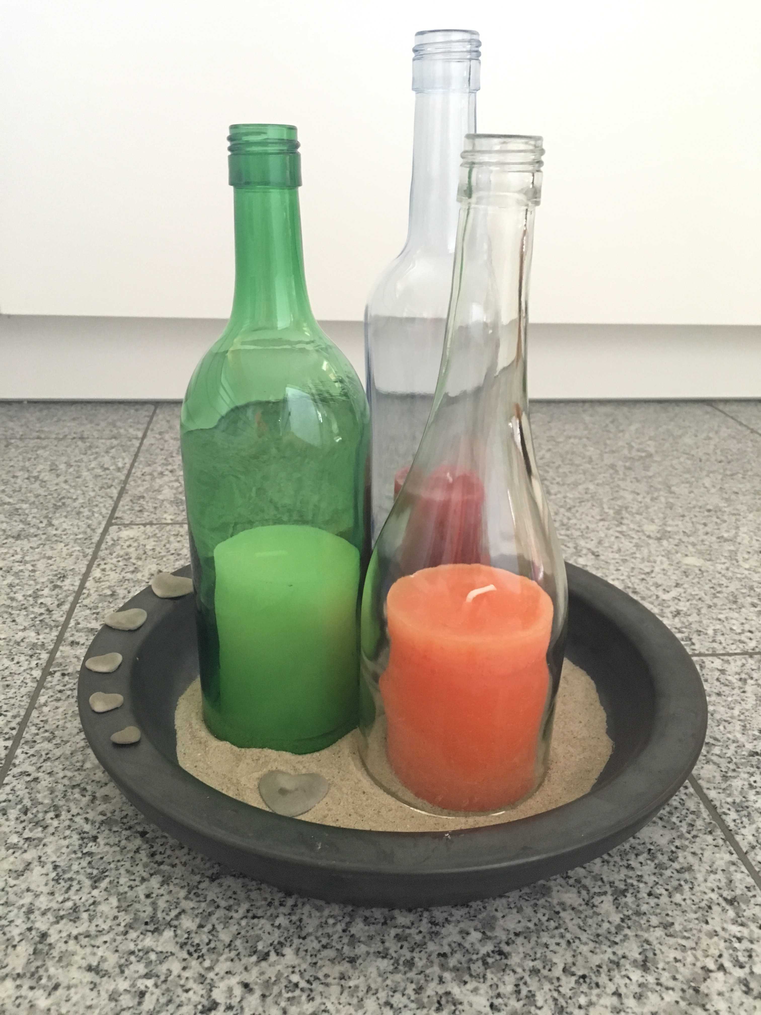 Auf einem Sandbett stehen Glasflaschen, in denen große Kerzen stehen.