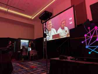 Videoleinwand neben Telefonbox, Personen auf der Leinwand und auf Stühlen davor