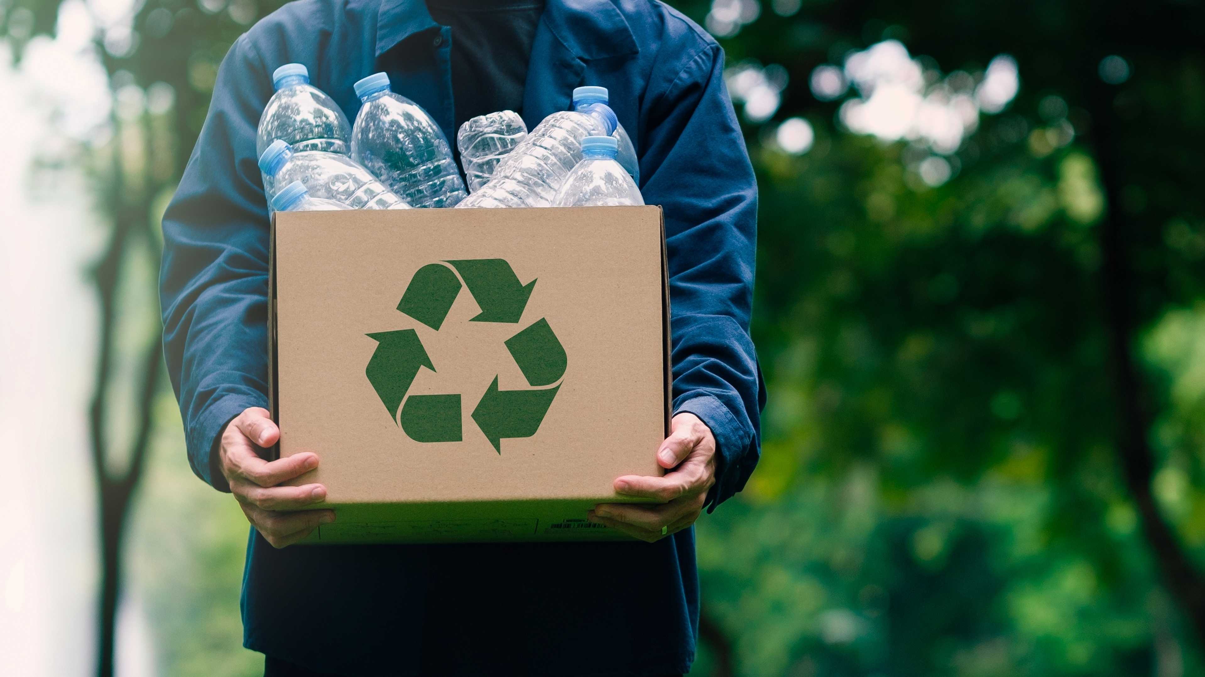 Ein Mann trägt eine Kiste mit leeren Plastikflaschen. Auf der Kiste ist ein Recycling-Zeichen.