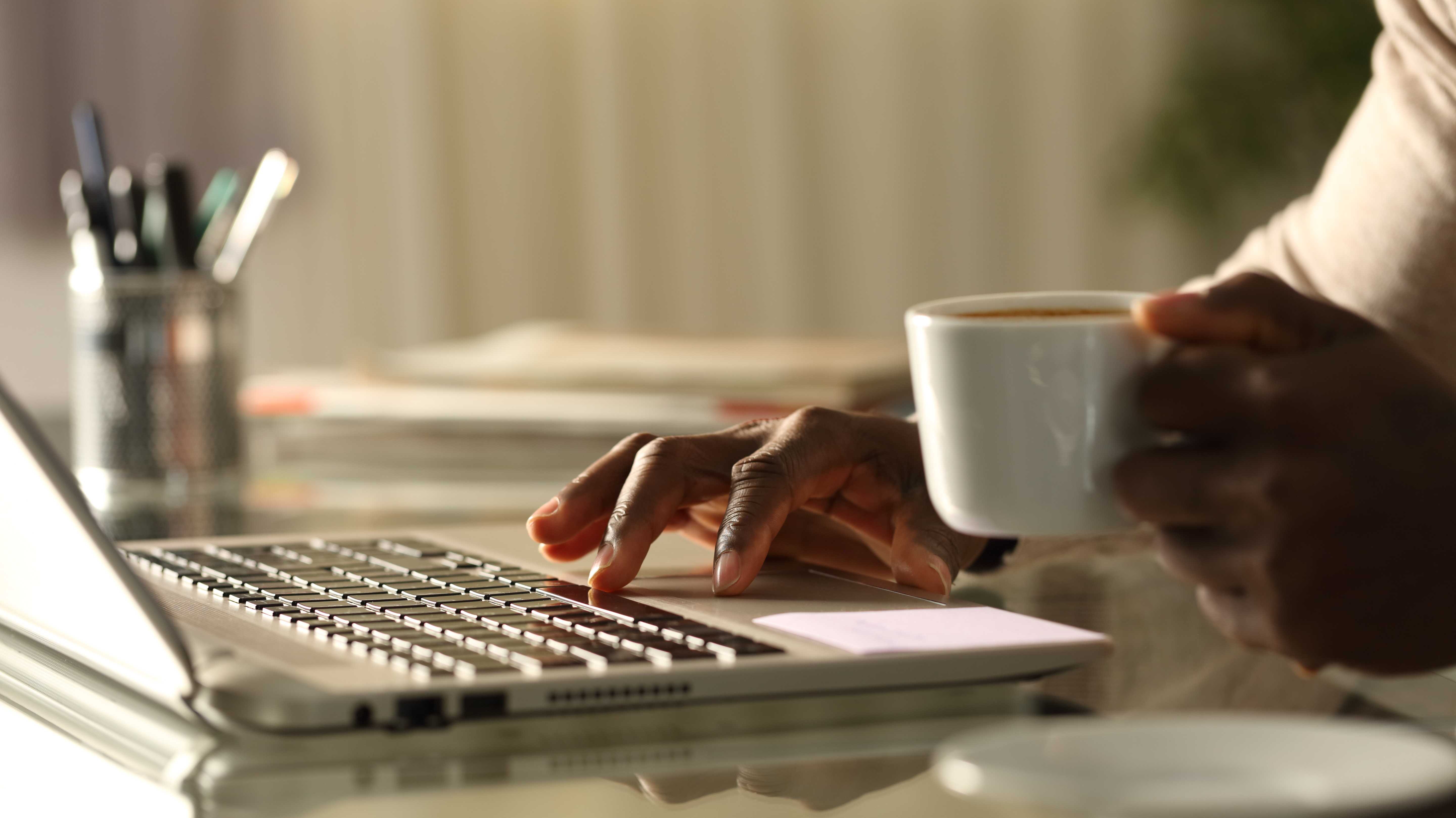 Person im am Schreibtisch, in einer Hand eine Kaffeetasse, die andere Hand an der Tastatur.