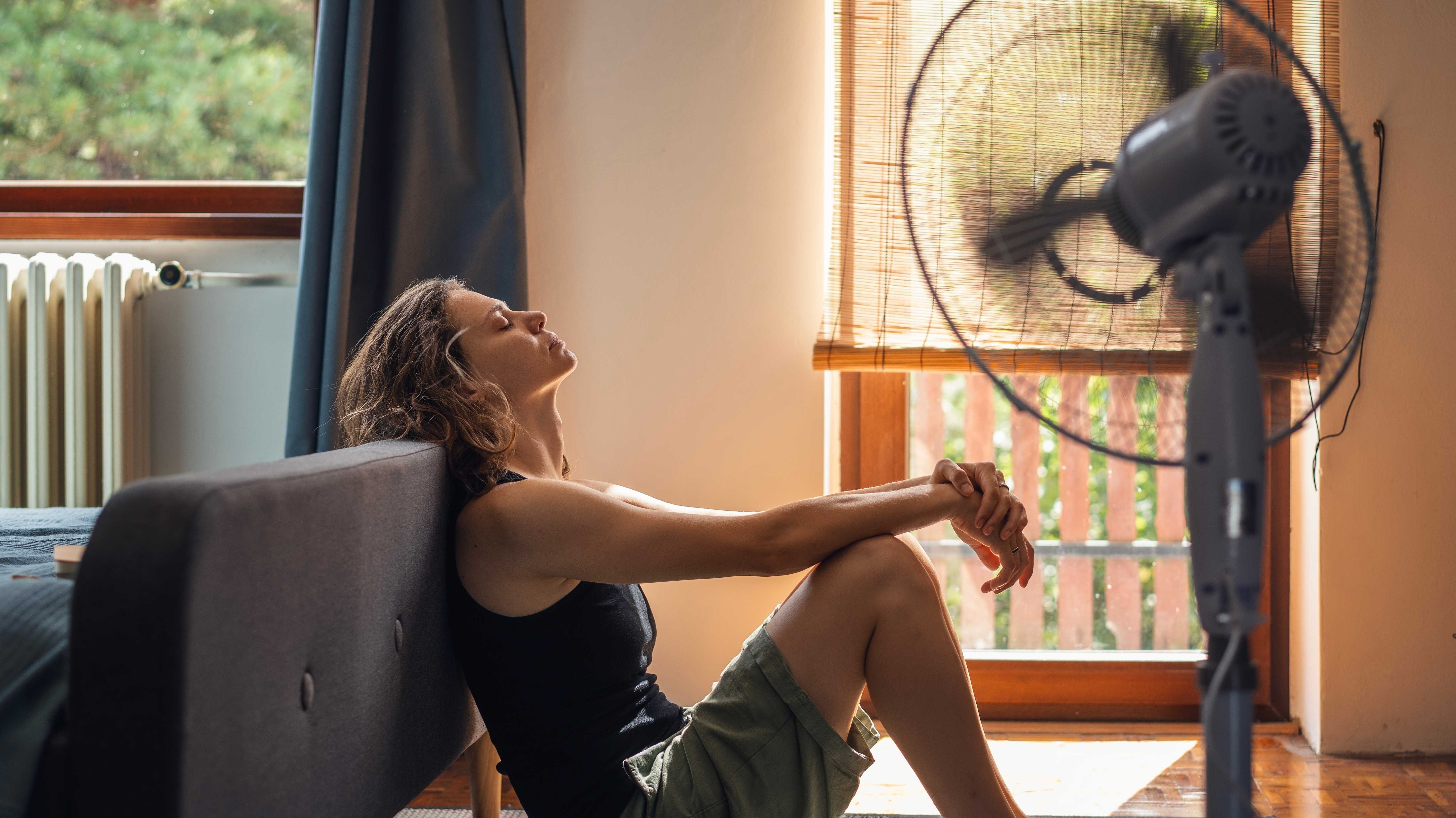 Junge Frau leidet unter Hitze, die zu Hause auf dem Boden vor einem Ventilator sitzt