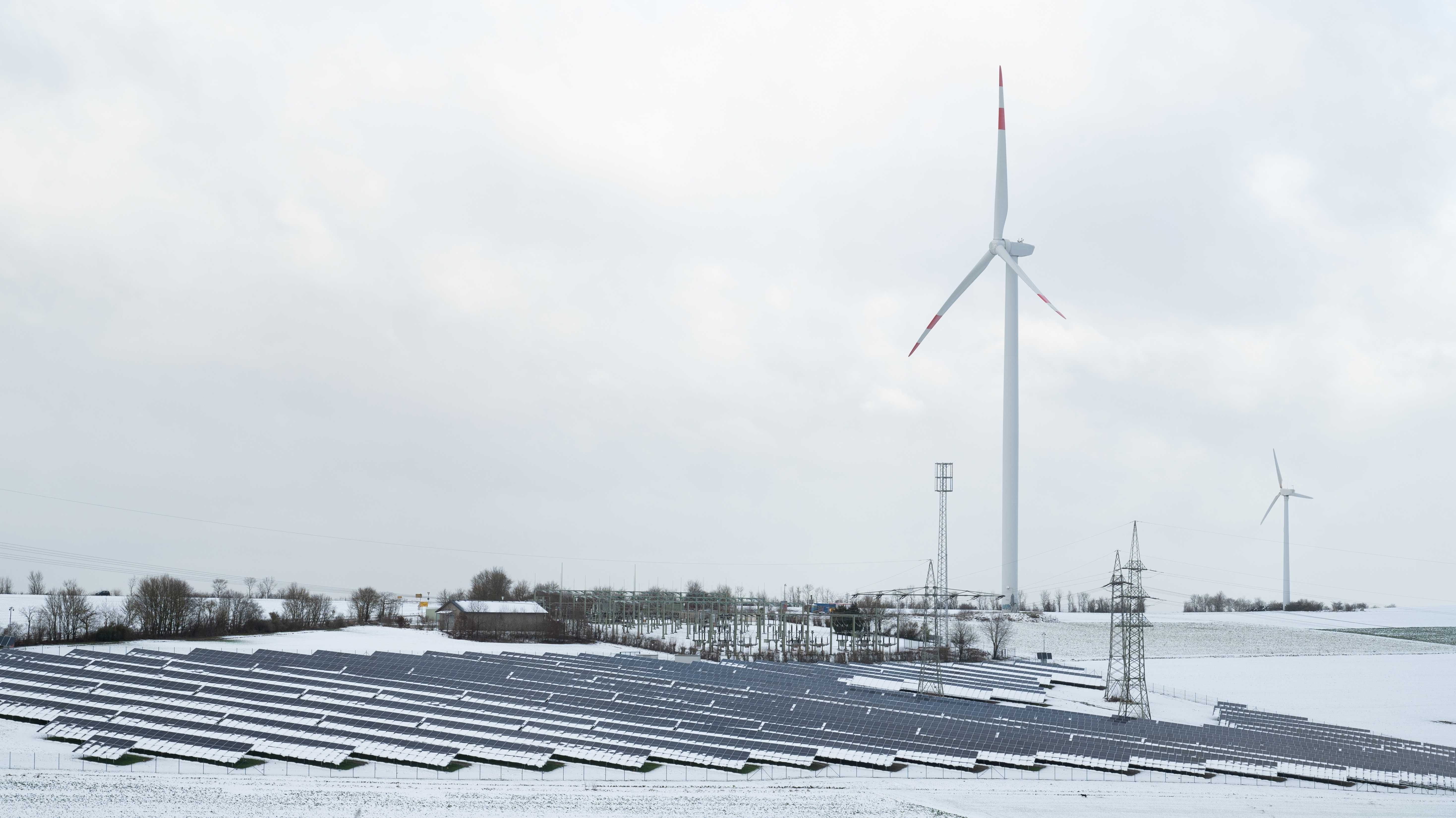 Stilstehende Windräder und Sonnenkollektoren in bewölkter schneebedeckter Landschaft.