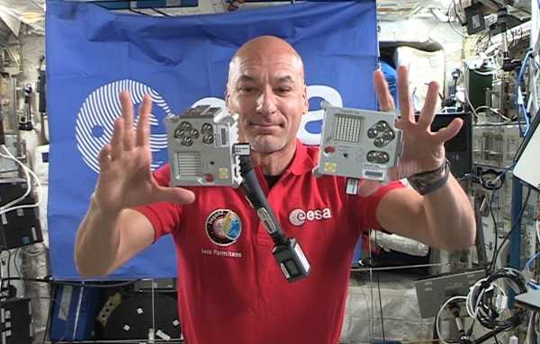In der Raumstation ISS fliegen zwei silberne Gehäuse, in denen Raspberry Pis eingebaut sind, vor einem Astronauten und der ESA-Flagge durch die Luft.