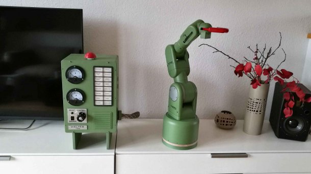 Ein grüner Roboter-Arm neben einem grünen Schaltgehäuse und einer Vase mit roten Blumen