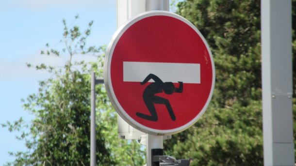 Verkehrszeichen "Ausfahrt einer Einbahnstraße" mit zusätzlich aufgemalter Figur, die quasi den weißen Querbalken auf der Schulter schleppt
