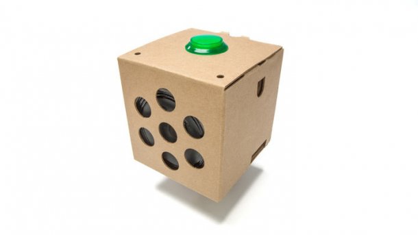 Ein brauner Pappwürfel mit grünem Button oben und Lautsprecher hinter großen Löchern an der Seite