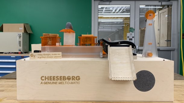 Maschine aus Holz: Oben hebt ein Arm Toastbrotscheiben und Käse. Ein Fließband transportiert sie in einen Sandwichgrill.