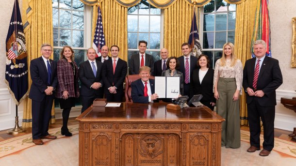 Trump hält sitzend ein Dokumentenmappe hoch hinter ihm steht etwa ein Dutzen Leute stramm und lächelt