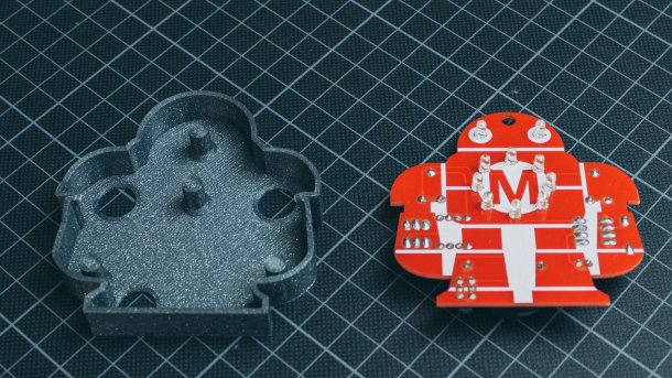Auf einer Millimetermatte liegen eine rote Platine in Form eines Roboters und daneben ein Gehäuse aus dem 3D-Drucker.