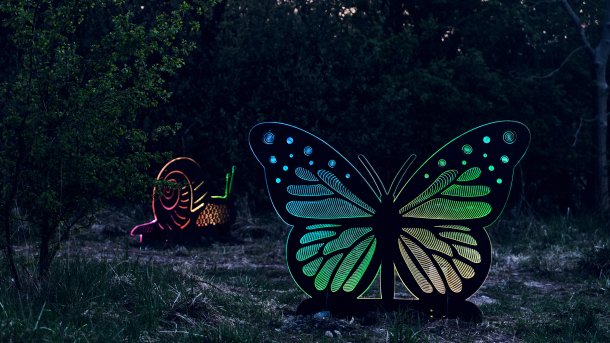 Eine überlebensgroße Schnecke und Schmetterling stehen als beleuchtete Metallskulpturen zwischen Sträuchern.