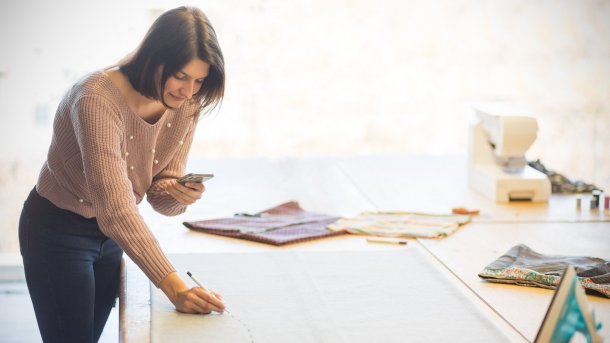 Eine junge Frau beugt sich über einen Tisch mit Stoff. Sie schaut auf ein Smartphone in der linken Hand und zeichnet mit der rechten Hand auf Stoff.
