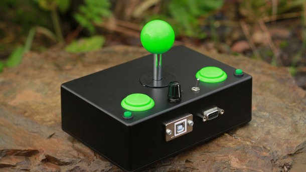 Ein schwarzes, rechteckiges Gehäuse aus Metall, aus dem zwei grüne Buttons, ein Kippschalter, ein Potentiometer und ein grüner Joystick ragen.