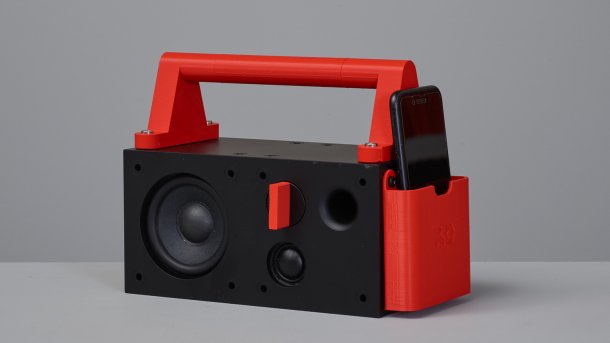 Eine schwarze, rechteckige Boombox mit rotem Griff und roter Seitenlasche für ein Handy.