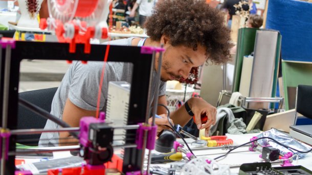 Ein junger schwarzer Mann sitzt hinter einem 3D-Drucker und lötet Bauteile