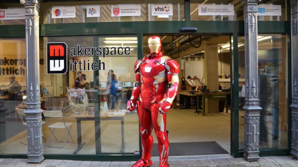 Iron Man in roter Rüstung vor einer Glaswand mit Aufschrift "Makerspace Wittlich". Dahinter sind Geräte eines Makerspaces zu erkennen.