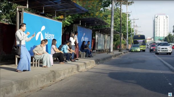 Personen warten an Bushaltestelle, dort "Telenor"-Werbeplakate
