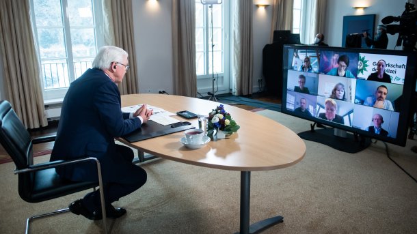 Mann sitzt alleine an ovalem Tisch, davor ein großer Bildschirm der mehrere Teilnehmer einer Videokonferenz zeigt
