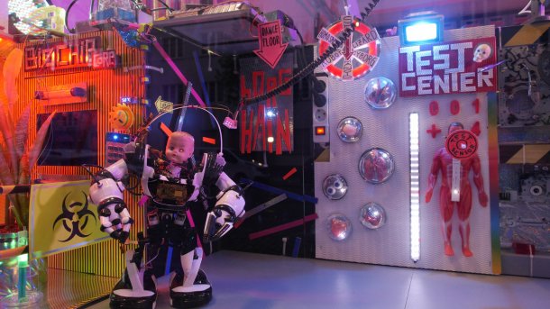 Ein Roboter mit Puppenkopf steht vor einer beleuchteten Kulisse mit "Häuserfronten" von Biochip Corp, Borghain und einem Testcenter.
