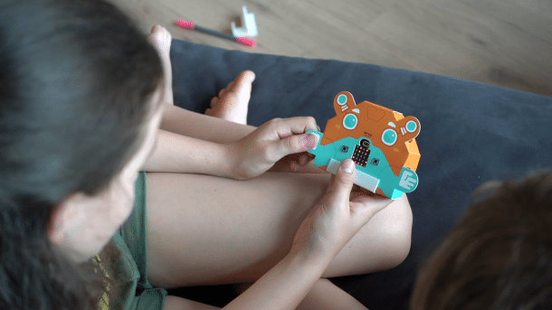 Zwei Kinder blicken auf einen bunten Papproboter mit LED-Matrix im Bauch.