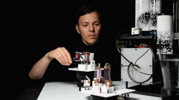 Ein weißer Mann schaut auf eine Maschine mit elektronischen Bauteilen und weißen Papierkügelchen in durchsichtigen Zylindern.
