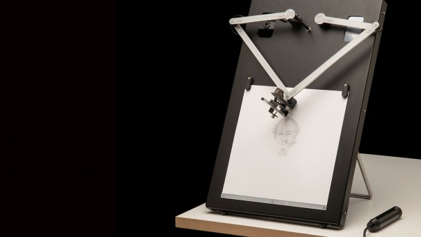 Auf einem Tisch steht ein schwarzer Kasten, auf dem ein weißes Papier eingeklemmt ist. Darüber halten zwei kleine Roboterarme einen Stift.