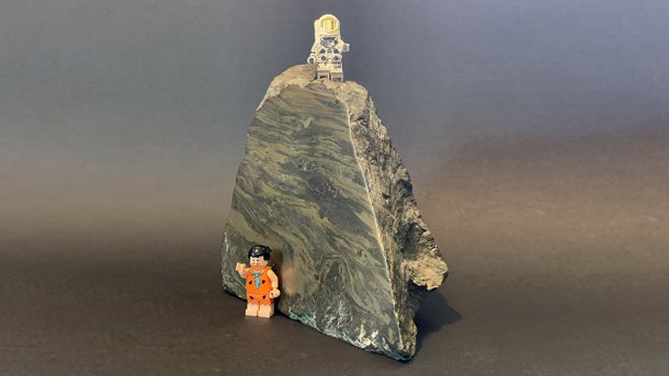 Ein Erzbrocken mit einer glattgeschliffenen Seitenfläche. Darauf und daneben zwei Legofiguren (Astronaut Neil Armstrong und Steinzeit-Cartoonfigur Fred Feuerstein).