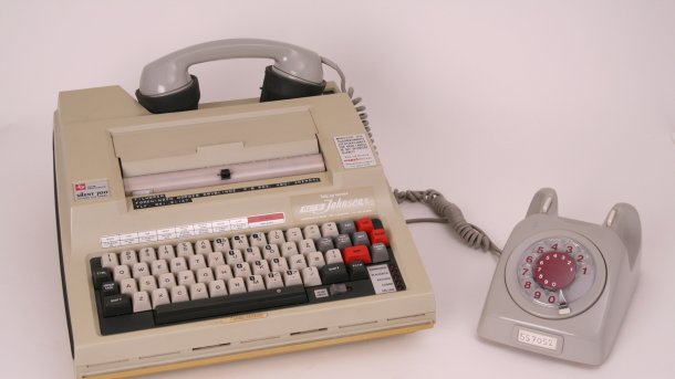 Ein beiger Kasten mit Tasten, der TI-Silent 700, daneben ein Wählscheibentelefon. Der Hörer des Telefons liegt oben auf dem Kasten auf Gummi-Anschlüssen.