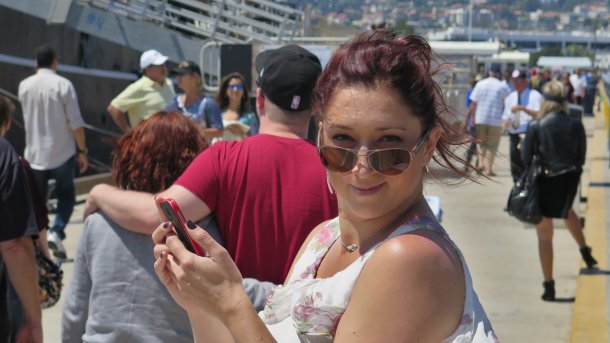 Eine Frau hält mit beiden Händen ein rotes Handy. Sie blickt über ihre Sonnenbrille hinweg in die Kamera. Im Hintergrund ein Pärchen und mehrere weitere unbeteiligte Menschen.