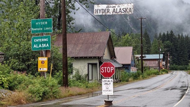 Grenzübergang von Kanada nach Hyder, Alaska - Stoppschild in der Straßenmitte, über der Straße hängt ein Schild "Welcome to Hyder, Alaska", links der regennassen Straße Nadelbäume und einige Häuser