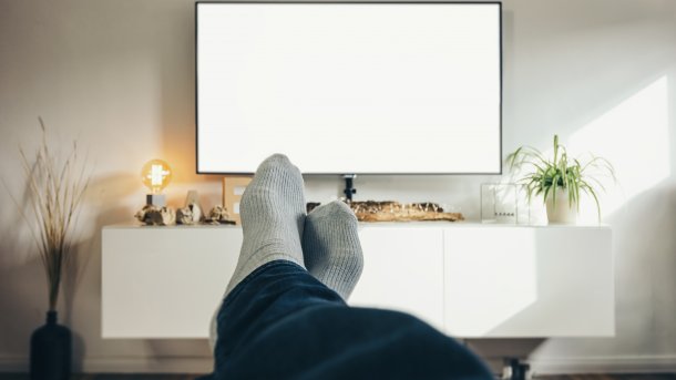 Die Beine hochgelegt, blickt eine weiß besockte Person auf einen hell erleuchteten Fernsehschirm